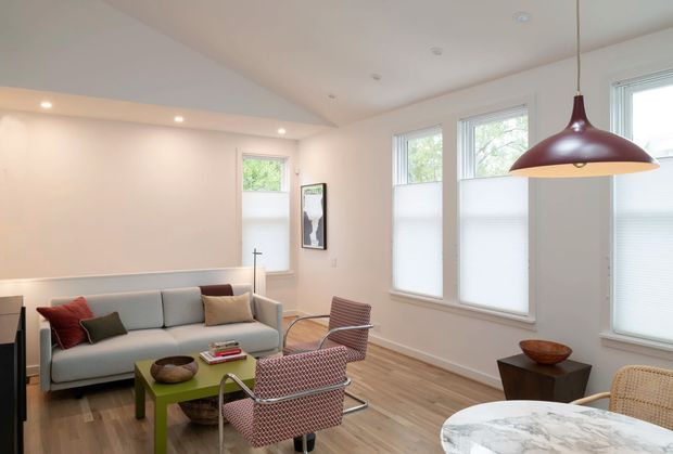 Bright contemporary living room with white walls, pale timber flooring, large windows fitted with translucent roller blinds, a light grey sofa, patterned lounge chairs, a green coffee table, and a round marble-topped dining table in the foreground. A large burgundy pendant light provides a bold accent, while recessed ceiling lights and abundant glazing create an airy feel.
