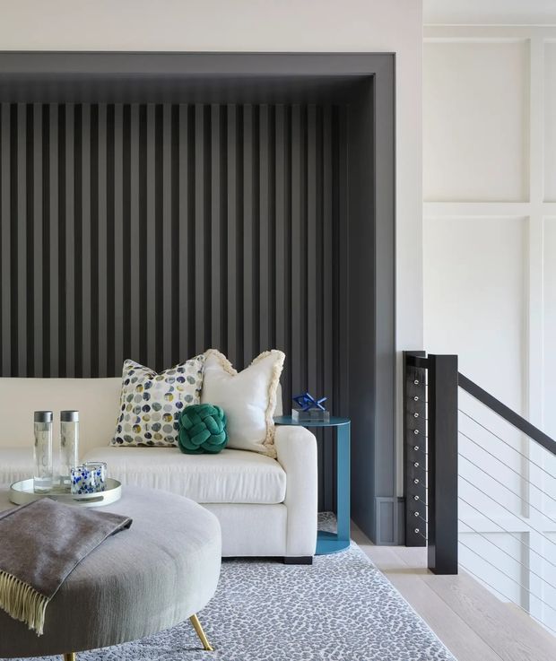 A refined contemporary living room with a pale upholstered sofa and rounded ottoman, layered cushions, a small blue side table, patterned rug, and a dark vertical fluted feature wall. A black-and-white open stair balustrade is visible at right, with white decorative wall panelling and light timber flooring.