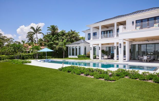 Bright exterior view of a large white two-storey residence with a low-pitched metal roof, covered balconies, extensive glazing, and a long rectangular swimming pool. The landscaped garden includes manicured lawn, clipped planting, tropical palms, and a poolside umbrella. The architecture has a refined resort-like appearance with strong horizontal lines and open outdoor living areas.