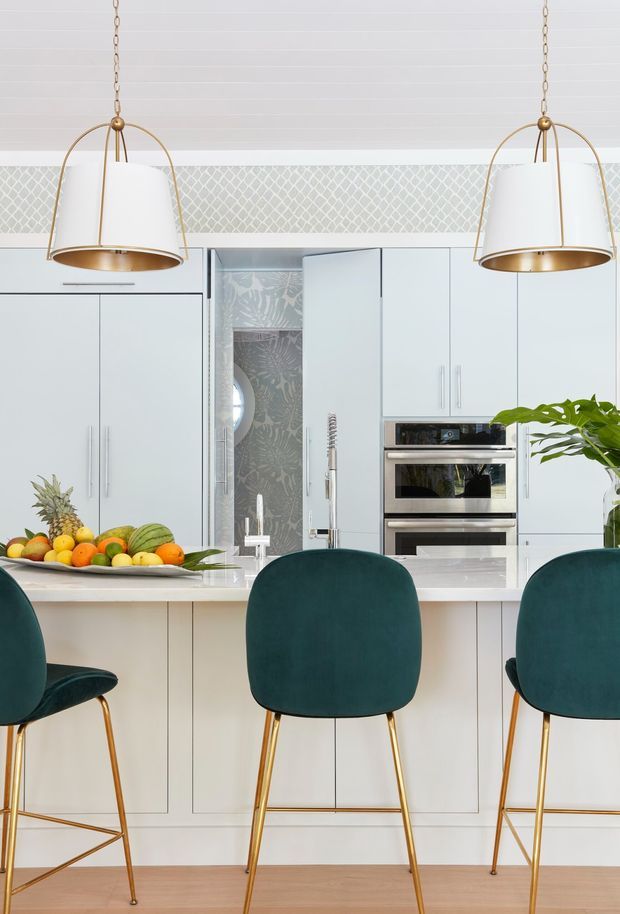 Bright contemporary kitchen with a large white island, pale flat-panel cabinetry, integrated stainless-steel appliances, and a patterned tiled backsplash. Three deep teal upholstered counter stools with slender brass-gold legs sit at the island, while oversized white and brass pendant lights hang above. Light timber flooring and fresh fruit add warmth and colour.