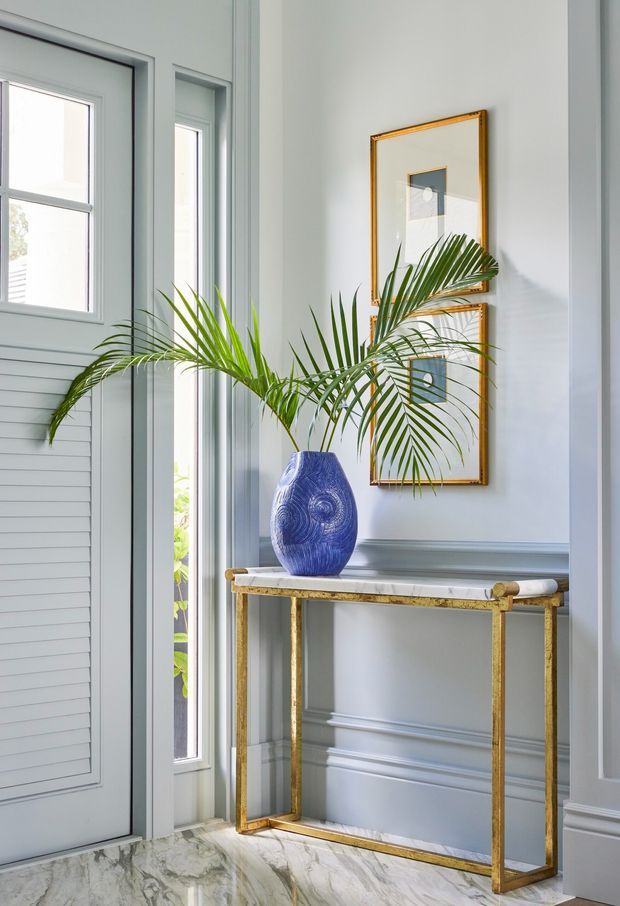 Bright, elegant entryway featuring pale blue-grey wall panelling and a louvred door beside tall windows. A slim gold-toned console table with a light stone top supports a sculptural blue vase and palm fronds, with two framed artworks above. The floor has a dramatic veined marble finish.