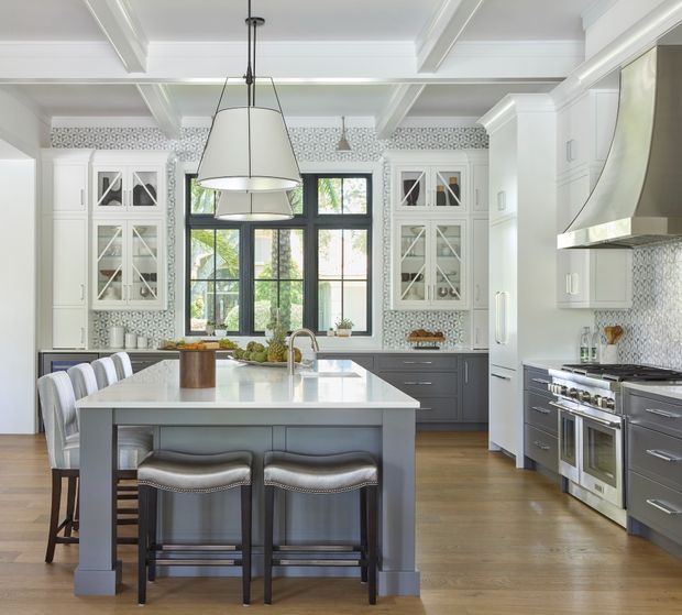 Bright, spacious traditional kitchen with a large central island, integrated seating, white coffered ceiling, white glass-front cabinetry, dark grey lower cabinets, patterned tile backsplash, stainless-steel appliances, and black-framed garden-facing windows. The space has a refined, classic look with abundant natural light.