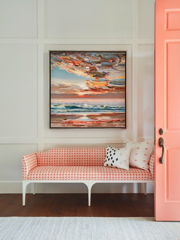A bright, carefully styled seating nook with a built-in or fitted patterned sofa against white panelled walls. A colourful seascape artwork hangs centrally above the sofa, while a coral-pink paneled door opens into the room from the right. The space has dark timber flooring, a pale textured rug, and decorative patterned cushions. The overall look is cheerful, coastal-inspired, and refined.