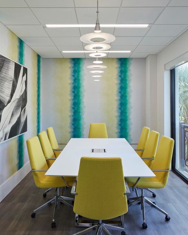 A bright contemporary meeting room with a long white rectangular conference table surrounded by yellow upholstered swivel chairs. The room features a vivid vertical abstract mural in teal, green, and pale yellow, dark wood-look flooring, a suspended row of circular pendant lights, a suspended-grid ceiling with linear fluorescent lighting, monochrome wall artwork, and full-height glazing on one side.