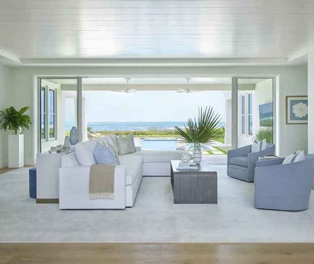 Bright coastal living room opening through wide glazed doors to a pool and ocean view. The room has pale upholstered sofas and armchairs, a low dark coffee table, light carpeting, white walls, a white paneled ceiling, ceiling fans, framed artwork, and indoor tropical plants. The setting feels airy, relaxed, and refined.