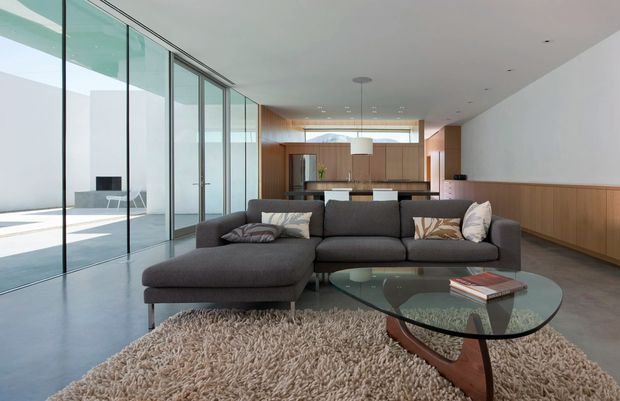 A spacious contemporary living room with a dark grey sectional sofa, glass coffee table, textured cream rug, and wood-panelled built-in cabinetry. Full-height glazed sliding doors open the room to a bright courtyard or exterior terrace, while a linear kitchen and dining area are visible beyond. The space has a clean, open-plan layout with warm timber detailing and abundant natural light.