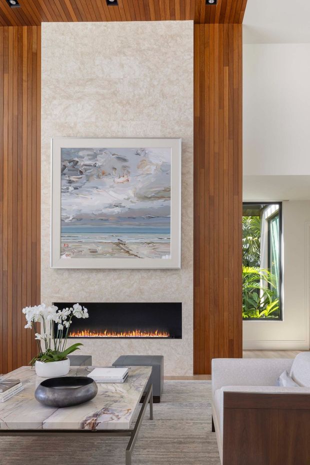 A refined contemporary living room centered on a linear built-in fireplace set into a pale stone feature wall. Warm vertical timber panelling extends across the walls and ceiling, while a framed coastal artwork hangs above the fire. A marble-topped coffee table, sculptural bowl, orchid arrangement, pale sofa, and neutral rug complete the calm, high-end interior. A large window reveals lush greenery outside.