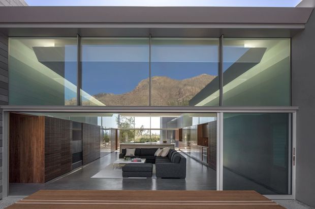 View into a contemporary living room through fully glazed sliding doors, with a dark sectional sofa, long open-plan sightlines, timber wall panels, and polished grey flooring. A dramatic mountain landscape is framed through the upper clerestory glazing, while the space features strong horizontal lines, exposed structural framing, and a high ceiling.