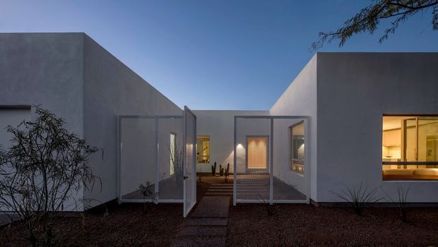 Exterior view of a low, minimalist modern residence at dusk, composed of smooth pale rendered volumes around a recessed entry courtyard. Mesh screen gates and a stepping-stone path lead toward the illuminated entrance, while a large window on the right reveals a warm-toned interior. Sparse desert-style landscaping and a deep blue sky reinforce the restrained architectural composition.