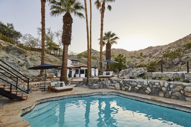 A resort-like outdoor pool area set within a rugged rocky desert landscape. A turquoise swimming pool occupies the foreground, surrounded by natural stone retaining walls, paved terraces, loungers, dark umbrellas, palm trees, and a small white building. The setting has a warm, rustic Mediterranean/desert character with dramatic rocky hills immediately beyond.