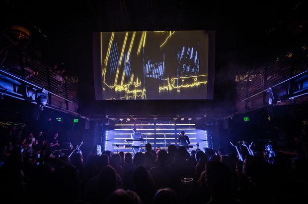 A dark, atmospheric live music venue with a crowded audience facing a raised performance stage. Blue and purple stage lighting, horizontal illuminated bars, DJ or electronic music equipment, side balcony structures, and a large overhead projection screen showing abstract yellow line imagery dominate the scene.