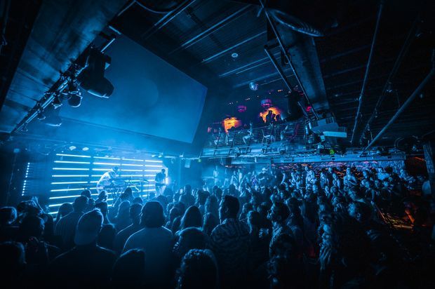 A dark, energetic nightclub or live music venue filled with a dense crowd. Blue stage lighting and horizontal LED light bars illuminate performers at the front, while a large projection screen, overhead rigging, speakers, and a raised balcony with additional people and colorful lights define the commercial entertainment interior.