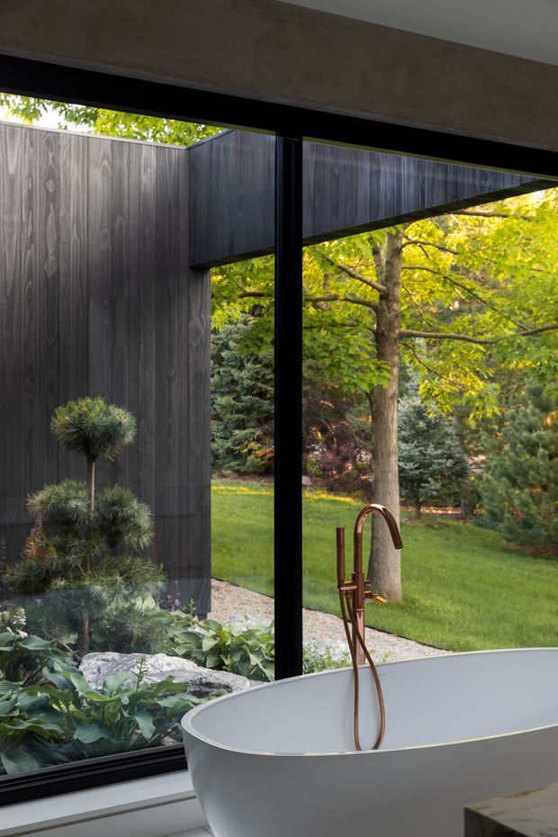 A refined bathroom interior centred on a freestanding white bathtub beside full-height black-framed glazing. The glazing looks onto a landscaped garden with mature trees, lawn, rocks, and planting. A dark vertical timber-clad privacy wall sits outside, while a sculptural copper-toned bath tap provides a warm accent. The image has a calm, nature-connected contemporary architectural character.