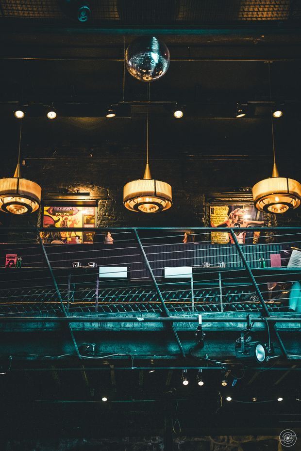 A dark, atmospheric commercial interior with exposed black-painted brick walls, a reflective disco ball, industrial track lighting, and three large pendant fixtures. A metal-railed mezzanine or balcony spans the space, with posters and signage on the rear wall and stage or venue equipment visible below. The image has a moody nightclub or live-music venue character with teal and warm amber lighting.