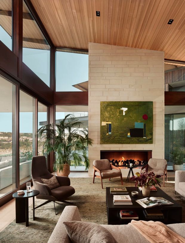 A sophisticated living room with a dramatic floor-to-ceiling glazed corner, dark timber window frames, a warm timber-lined sloping ceiling, and a pale brick fireplace wall. The fireplace is topped with a large abstract artwork, while layered lounge seating, a dark coffee table, indoor palms, and neutral textured furnishings create a refined, warm atmosphere. A view of the surrounding landscape is visible through the glazing.