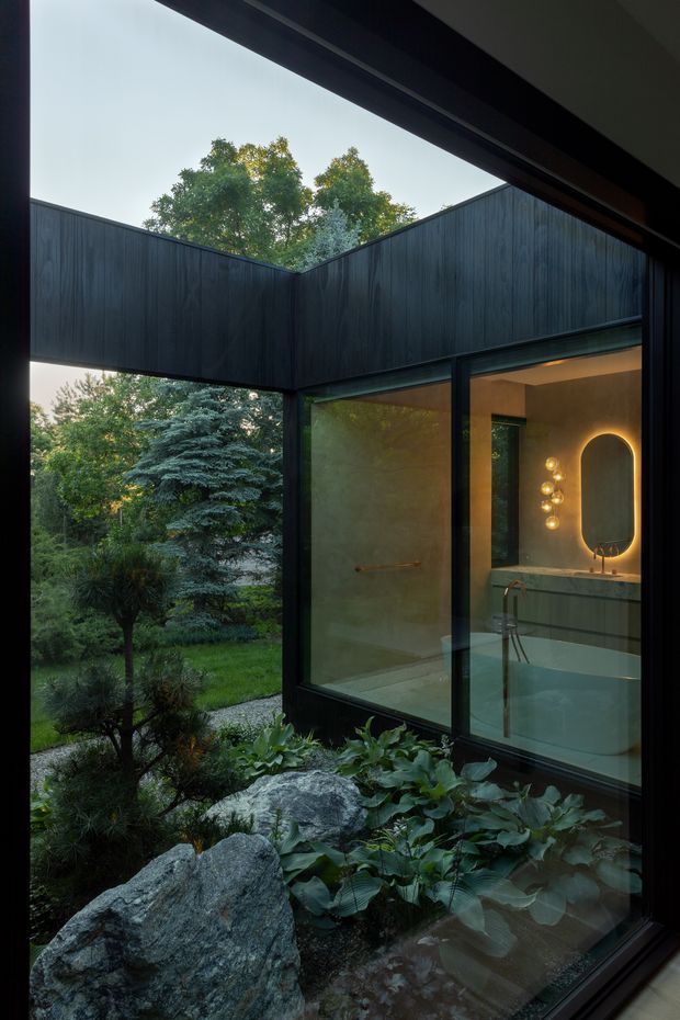 A contemporary bathroom viewed through full-height glazing beside a landscaped courtyard garden. The composition features dark timber-clad exterior walls, black-framed glass, a freestanding bathtub, stone vanity, illuminated oval mirror, and warm decorative wall lighting. Large rocks, lush planting, and mature trees form a private garden immediately outside, creating a strong indoor–outdoor connection.