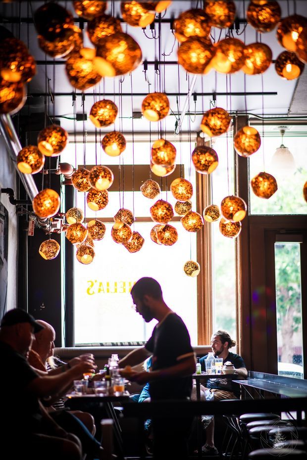 Interior photograph of a lively restaurant or café dining space, viewed toward large street-facing windows and a dark timber-framed entrance. The ceiling is densely covered with many suspended spherical pendant lights in warm amber and copper tones, creating a dramatic canopy above diners. Tables, chairs, patrons, and a server occupy the lower half of the image. The space has an industrial-style exposed ceiling and dark structural elements, with strong daylight contrasting against the warm lighting.