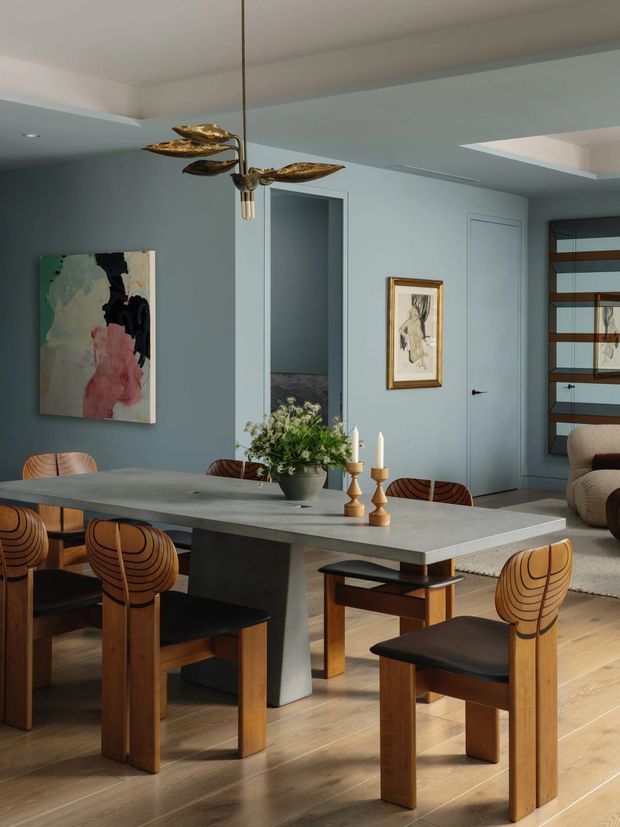 A refined dining area with a large grey stone or concrete dining table surrounded by sculptural timber and black upholstered chairs. The room features muted blue-grey walls, light timber flooring, a linear shelving and doorway composition, framed artwork, a contemporary sculptural pendant light, and a glimpse of an adjacent living area. The styling blends contemporary and mid-century influences with warm timber accents.