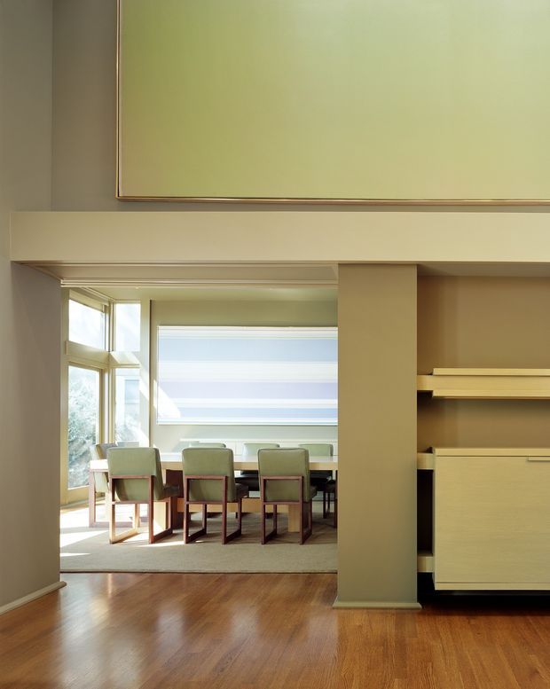 View through a wide interior opening into a bright dining area, with a rectangular dining table and upholstered chairs arranged in front of large windows. The foreground has warm timber flooring and a built-in wall unit or shelving on the right, while the room features muted beige-green walls, substantial architectural trim, and layered horizontal ceiling and wall details.