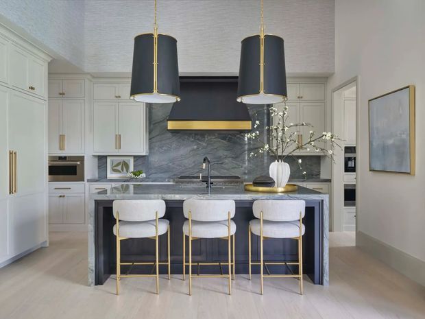 A refined contemporary kitchen with a large dark stone island, three upholstered bar stools with gold-toned frames, white built-in cabinetry, and a matching dark stone splashback. Two oversized black-and-brass pendant lights hang above the island, while integrated appliances and understated artwork complete the polished, neutral-toned scheme.