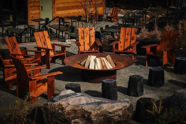 An outdoor communal seating area with multiple rustic timber Adirondack-style chairs arranged around a large circular steel fire pit filled with split logs. Rough stone stools and boulders form additional seating, with gravel paving, timber-clad structures, picnic-style tables, and sparse landscaping creating a rugged, natural setting.