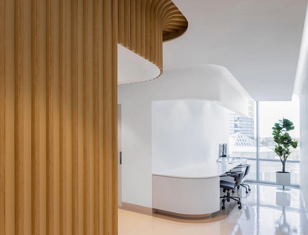 A bright contemporary office reception or workstation area with a sculptural curved white built-in desk, glossy pale flooring, floor-to-ceiling glazing, and a dramatic timber slatted feature wall and rounded overhead timber detail. Office chairs, a monitor, and a potted plant are visible, with an urban building seen through the windows.