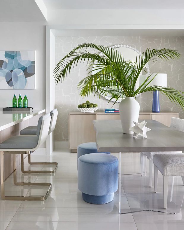 Bright contemporary dining area with a large rectangular dining table, upholstered dining chairs, blue cylindrical stools, and a tall potted palm. The room features glossy light flooring, pale built-in cabinetry, a textured light stone-pattern feature wall, white architectural trim, and a small adjacent breakfast bar with stools. Blue and grey accents create a fresh, airy interior.
