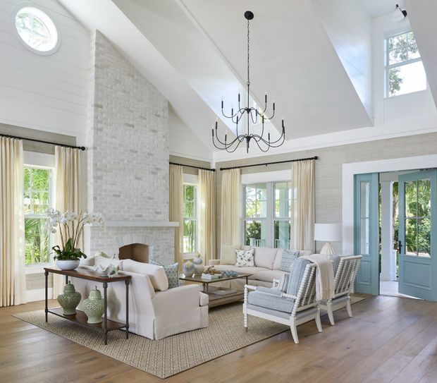 Bright, spacious living room with a vaulted white shiplap ceiling, white-painted brick fireplace, tall multi-pane windows, pale blue French doors, and a large black multi-arm chandelier. The room is furnished with light upholstered sofas and armchairs, a neutral textured rug, a timber coffee table, and a console decorated with ceramics and greenery. The overall look is refined, airy, and coastal-traditional.