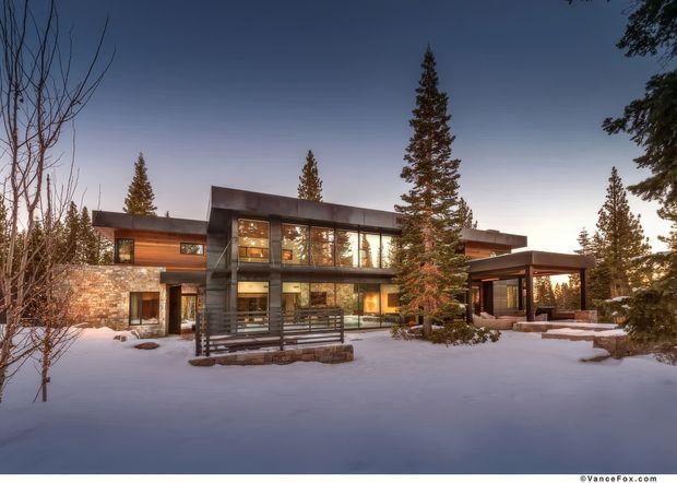 A contemporary multi-level mountain residence photographed in a snowy wooded setting at dusk. The home features broad floor-to-ceiling glazing, dark horizontal roof planes, warm stone masonry, timber and dark metal details, and a covered entrance or outdoor living pavilion. Snow-covered ground and mature evergreen trees surround the building.