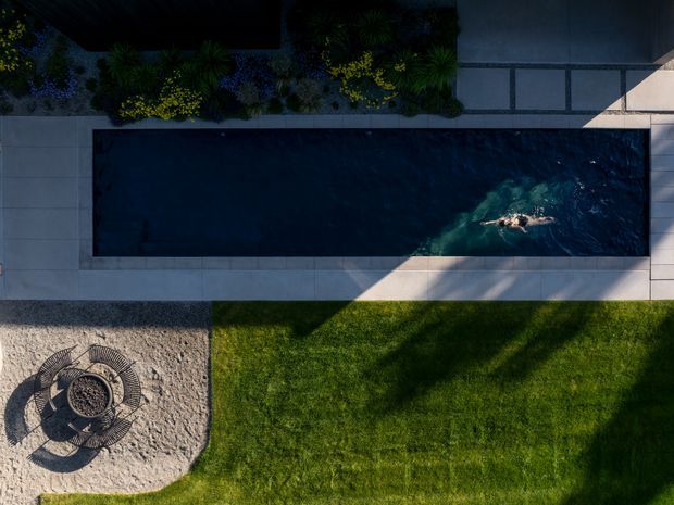Aerial view of a contemporary landscaped backyard centred on a long rectangular swimming pool. Pale paved terraces border the pool, with dense ornamental planting along one edge, a neatly maintained lawn below, and a circular outdoor fire pit or seating feature set into a pale gravel area. Strong afternoon shadows create a graphic composition.