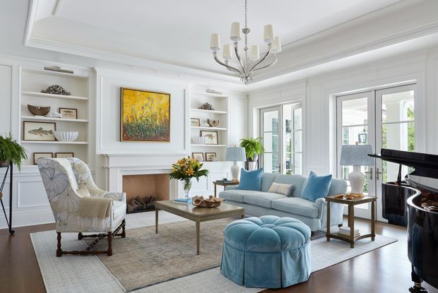 Bright, elegant living room with white paneled walls, built-in shelving, a classical fireplace mantel, large French doors, and a central chandelier. The room features a pale blue sofa and round tufted ottoman, patterned armchairs, a glass-topped coffee table, a neutral area rug, and a black grand piano. The overall look is refined and traditionally inspired with fresh coastal blue accents.