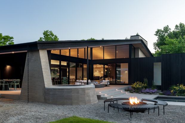 Modern single-storey residence with a dramatic flat roof, dark vertical cladding, extensive floor-to-ceiling glazing, and a projecting covered outdoor entertaining area. The landscaped courtyard includes a circular outdoor fire pit, gravel paving, built-in curved masonry feature, low planting, and illuminated steps. Warm interior lighting is visible through the glazing, with an outdoor dining area at the left.