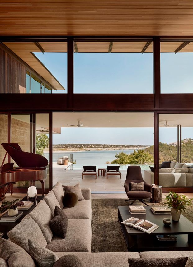 A refined living room with expansive floor-to-ceiling glazing opening to a waterfront view and outdoor terrace. The interior features warm timber-lined ceilings, dark timber window and door frames, a large neutral sectional sofa, sculptural lounge chairs, a black grand piano, and a low dark coffee table. The room is softly furnished and visually connected to an outdoor pool or terrace overlooking the water.