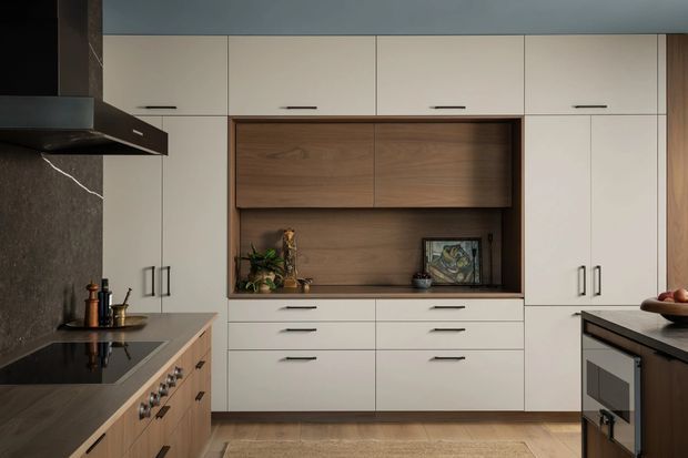 A contemporary kitchen with full-height off-white cabinetry, warm timber display and storage joinery, dark stone-look splashback, timber island cabinetry, integrated appliances, and black hardware. The composition is clean and refined with warm, muted tones.