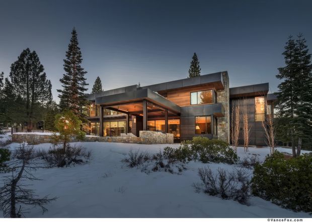 Exterior dusk photograph of a contemporary two-storey home set in a snowy, forested landscape. The house has strong horizontal flat-roofed forms, dark panelled cladding, extensive glazing, warm interior lighting, a projecting covered entry or terrace, and a low stone retaining wall.