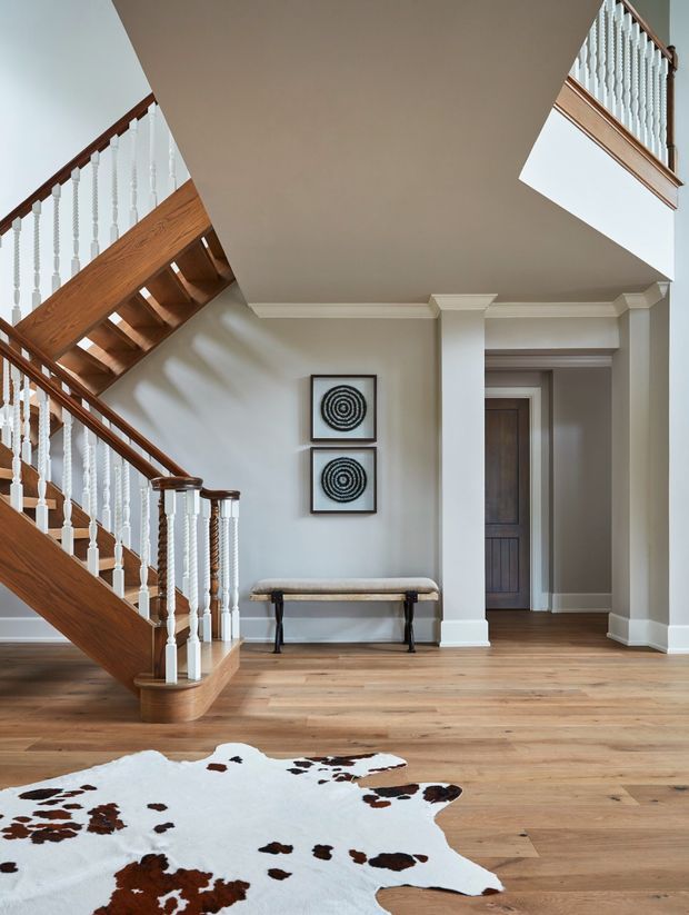 A bright, traditional residential entry hall dominated by a timber staircase with turned white balusters and dark wood handrails. The space has wide-plank timber flooring, pale painted walls, substantial white moulded trim and columns, a simple upholstered bench, framed artwork, and a cowhide rug. A hallway and doorway extend beyond the stair.