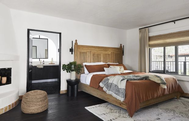 A warmly styled bedroom with a large woven rattan bed featuring a tall cane headboard and matching textured frame. The room has white walls and ceiling, dark wood flooring, a patterned area rug, layered neutral and rust-brown bedding, a woven round ottoman, a black pedestal side table, and a potted plant. A black-framed doorway opens to a bathroom with a dark vanity, while a large window with woven blinds and curtains provides natural light.