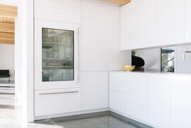 Bright minimalist kitchen with full-height white handleless cabinetry, an integrated glass-front storage unit, mirrored backsplash, and a grey polished concrete floor. A timber-lined ceiling and adjacent timber-lined space are visible through the opening at left, while a bowl and black decorative object sit on the benchtop.