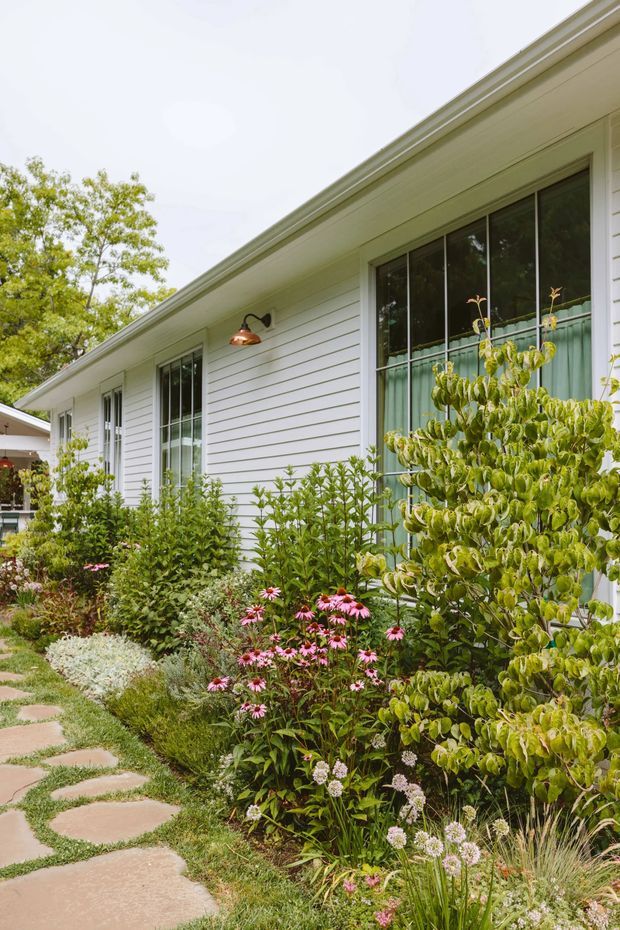 Exterior view of a low-rise house with white horizontal weatherboard cladding, a shallow pitched roof with broad eaves, tall dark-framed windows, and a copper wall-mounted light. A lush cottage-style garden with flowering plants borders an irregular stepping-stone path leading toward the entrance.