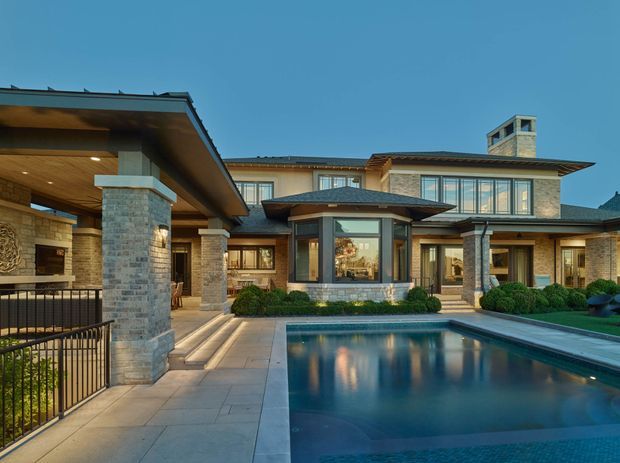 A large, sophisticated residential home arranged around a central rectangular swimming pool and paved courtyard. The two-storey facade combines pale stone and brick masonry, dark timber or metal rooflines, extensive glazed windows, covered outdoor entertaining areas, and substantial square columns. A pool pavilion or projecting bay forms a focal point at the centre, while landscaped hedges and formal planting soften the paved setting. The image is photographed externally at dusk, with warm interior and landscape lighting creating reflections across the pool.