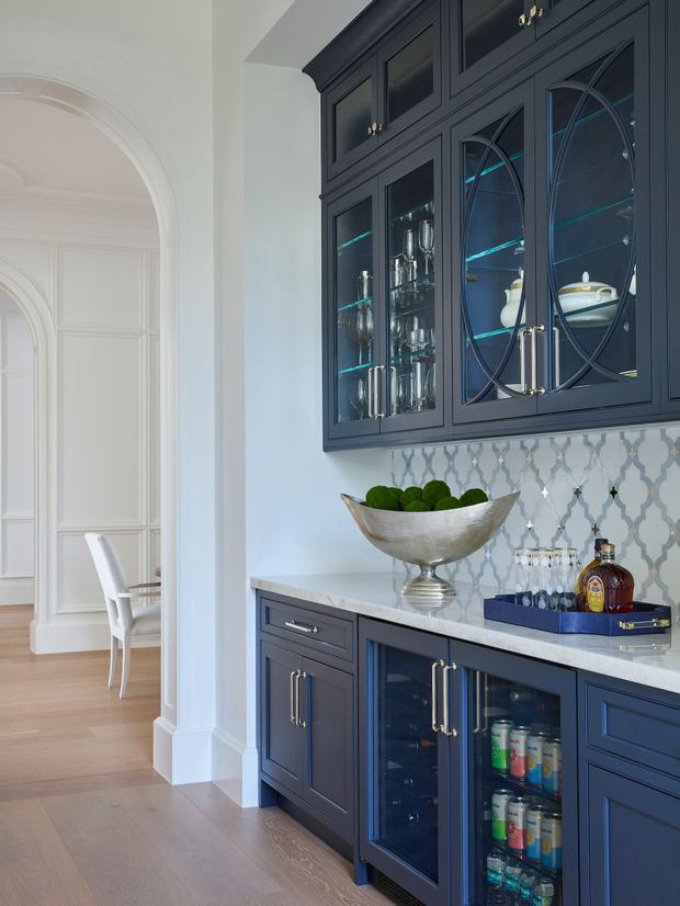 A refined built-in home bar or drinks station with deep navy cabinetry, glass-front upper cupboards displaying glassware and serving pieces, a pale stone benchtop, and a patterned light tile backsplash. The lower cabinetry includes a glass-front beverage refrigerator. An ornate white-trimmed archway opens toward a formal dining area with paneled walls and timber flooring.