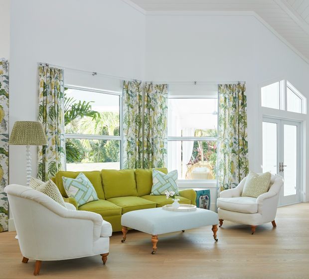 Bright, airy living room with white walls and trim, pale timber flooring, large windows, and glazed French doors. A lime-green sofa with patterned cushions is paired with cream upholstered armchairs and a matching tufted ottoman, while botanical curtains and tropical greenery outside add a fresh, relaxed character.