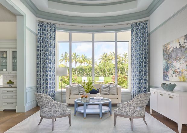 Bright, refined sitting room with a curved bay window overlooking tropical landscaping. The room features pale blue-grey wall panelling and layered ceiling mouldings, patterned blue-and-white curtains, a pair of upholstered lounge chairs, a light sofa, a central coffee table, a pale sideboard, and an area rug.