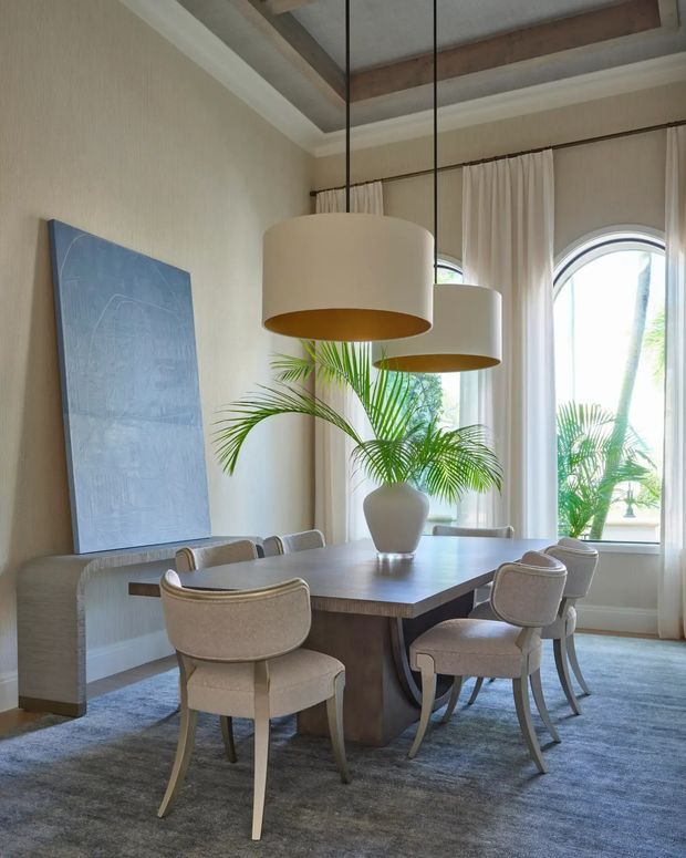 Elegant dining room with a substantial dark wood dining table, upholstered curved-back dining chairs, a large textured wall artwork, and two oversized cream pendant lights. Tall arched windows with sheer curtains bring in abundant daylight, while a potted palm and soft neutral finishes create a refined, airy atmosphere. A recessed timber ceiling detail adds architectural interest.