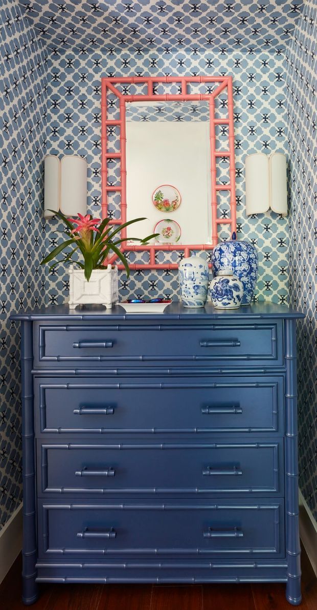 A compact, highly decorated interior vignette featuring a tall navy-blue painted chest of drawers with raised trim and metal pulls. The chest is topped with blue-and-white ceramic vessels, a white planter with a tropical plant, and small decorative objects. Behind it, blue patterned wall tiles or wallpaper cover the walls, with a coral-pink bamboo-style framed mirror and matching cream wall sconces. The overall look is colourful, eclectic, and vintage-inspired.