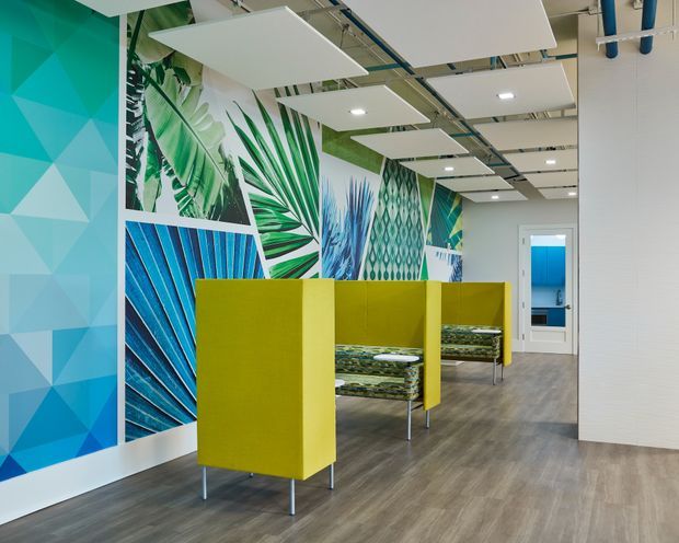 Bright contemporary commercial interior, likely an office or workplace breakout area, featuring vivid tropical and geometric wall graphics, lime-green acoustic privacy booths with patterned upholstered seating, grey wood-look flooring, white walls, and suspended white acoustic ceiling panels with exposed services. A blue-toned room is visible through the doorway at the rear.