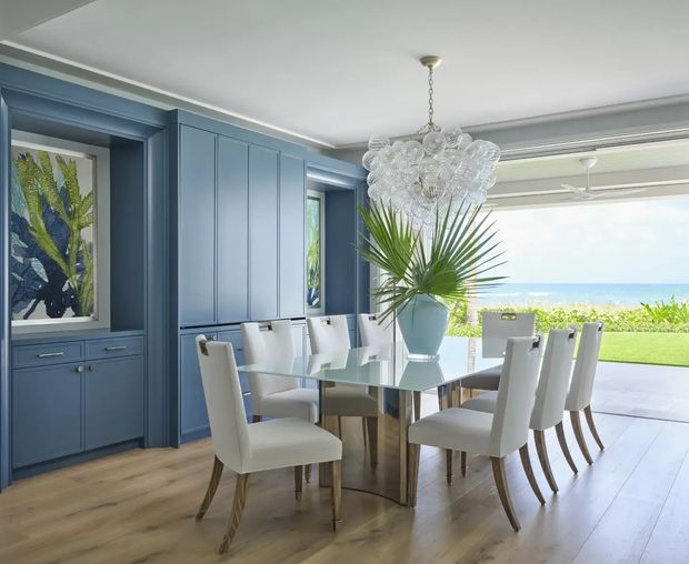 Elegant coastal dining room with a glass dining table and upholstered chairs, surrounded by deep blue built-in cabinetry and wall panelling. A sculptural glass-bubble chandelier hangs above the table, while large open glazing frames a lawn and ocean view. Warm timber flooring and tropical greenery add contrast to the refined blue-and-white interior.