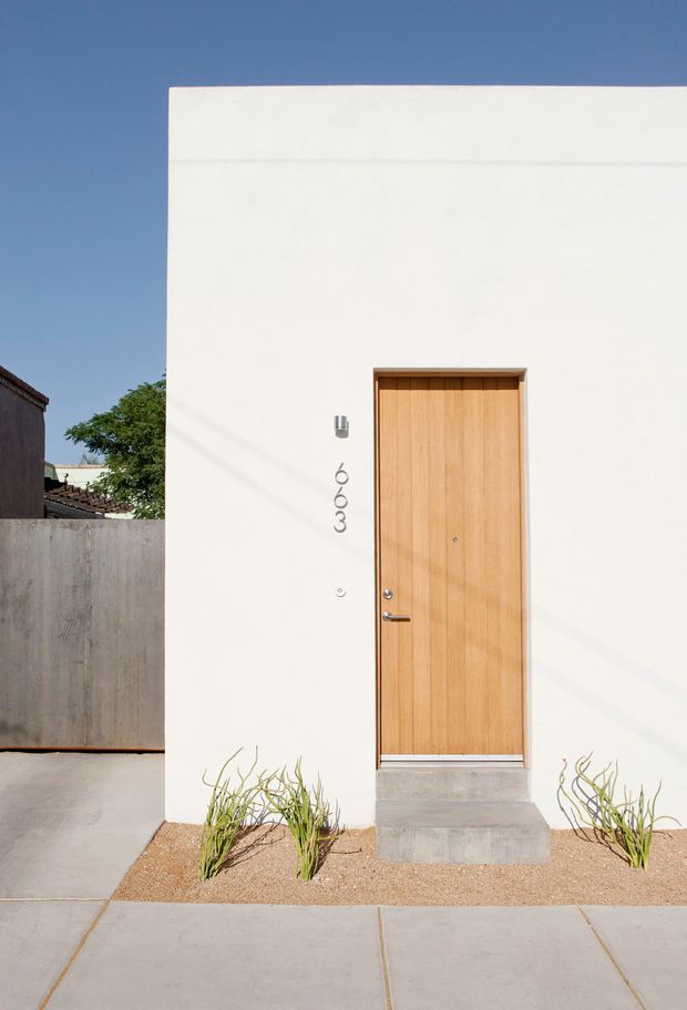 Minimalist exterior view of a small modern residence entrance. A smooth white rendered facade with a flush natural timber front door, simple metal door hardware, vertical house numbering, and a concrete entry step faces a paved sidewalk. Sparse drought-tolerant planting sits in a gravel bed, with neighbouring structures partially visible at the edges.
