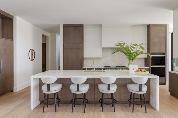 A bright, spacious contemporary kitchen featuring a large white waterfall island with seating for four, integrated sink and cooktop, dark timber cabinetry, pale timber flooring, white walls, and a stainless-steel refrigerator and appliances. A tall indoor palm adds greenery, while a hallway and adjacent cabinetry are visible in the background.
