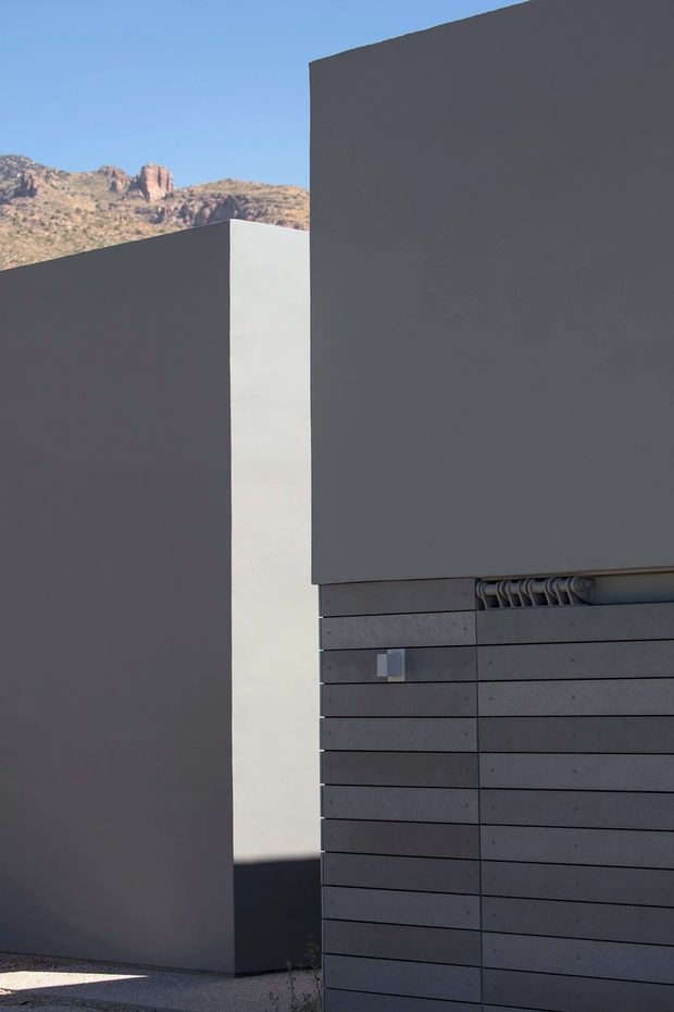 Minimalist exterior view of a sharply geometric building composed of dark charcoal rendered walls and horizontal dark cladding panels. A narrow white vertical wall creates a strong contrast, while rugged sunlit mountains are visible in the background beneath a clear blue sky. The image emphasizes abstract massing, clean lines, and concealed or integrated garage-door detailing.